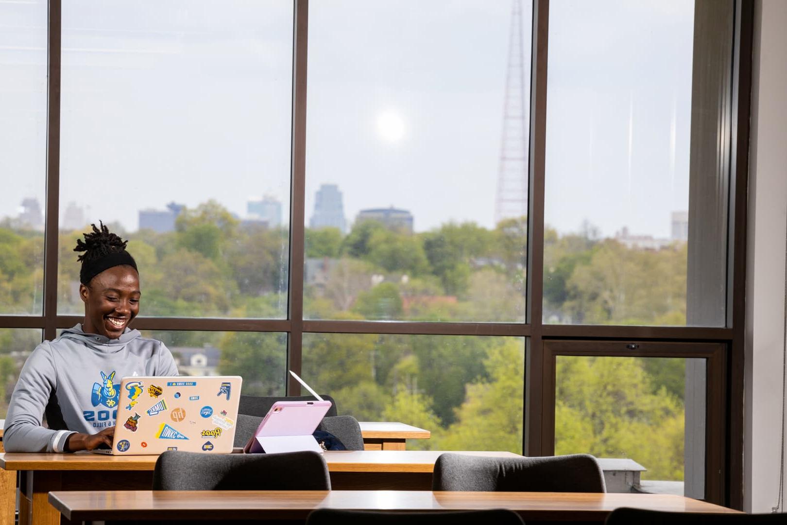 戴着发带的年轻女子坐在图书馆的一张桌子旁，用笔记本电脑和平板电脑工作，她身后的窗户可以看到堪萨斯城的天际线